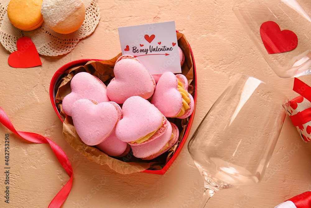 Box with tasty heart-shaped macaroons, glasses and greeting card on beige background