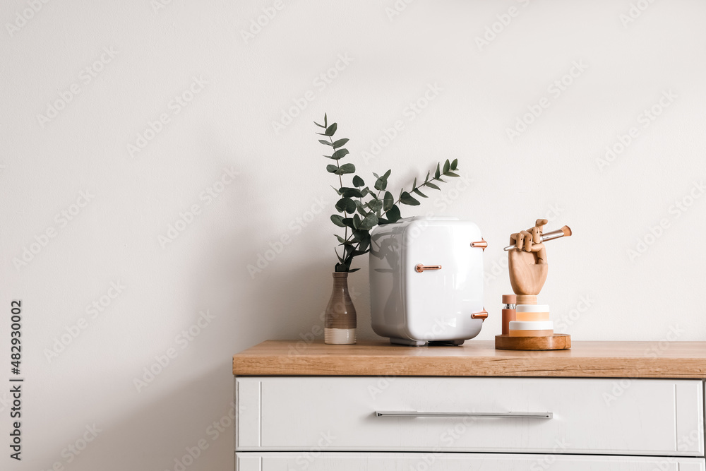 Small cosmetic refrigerator, vase with eucalyptus and wooden hand on table near light wall