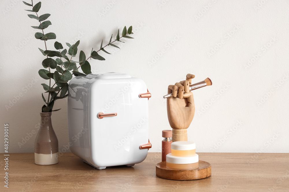 Small cosmetic refrigerator, vase with eucalyptus and wooden hand on table near light wall