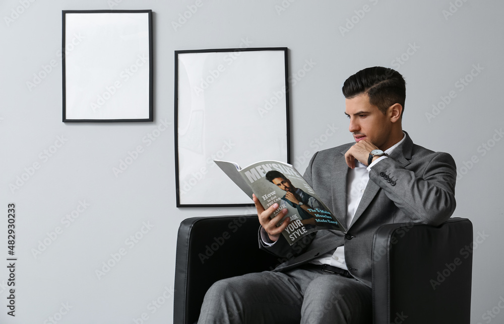 Fashionable man reading magazine in comfortable armchair near grey wall