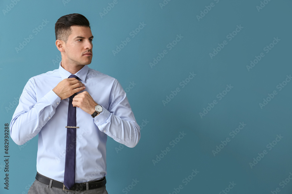 Young handsome man in formal clothes looking aside and adjusting tie on color background