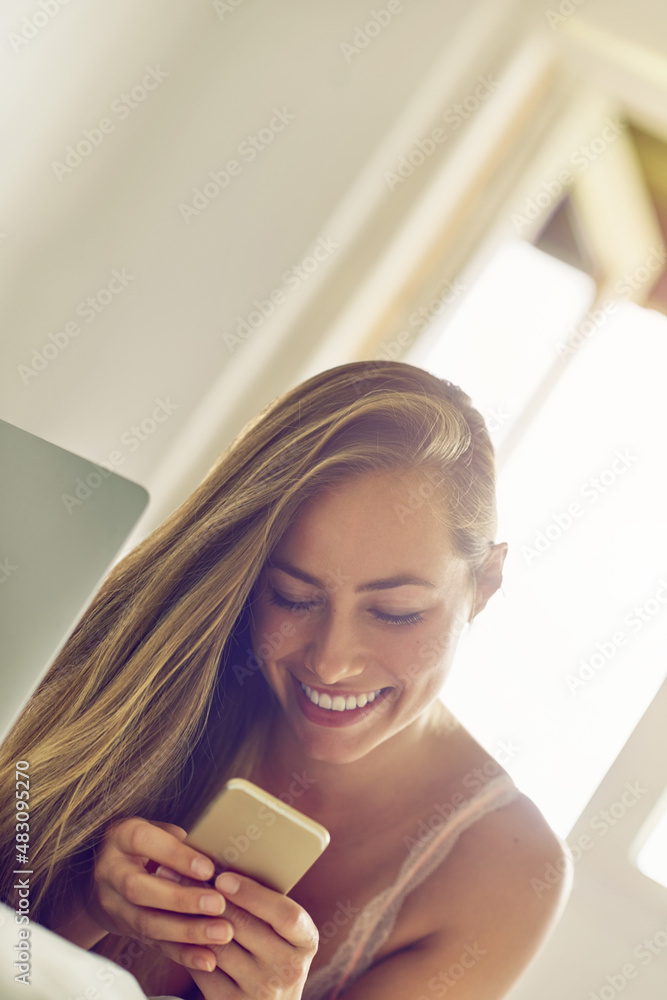 Sending a flirty text message. Shot of a young woman using a laptop and phone in the morning at home