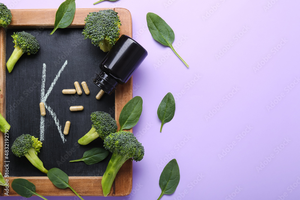 Chalkboard with letter K, bottle of pills, broccoli and spinach leaves on lilac background, closeup