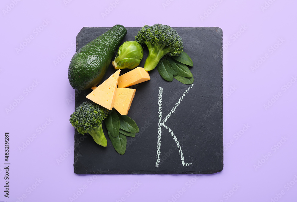 Slate board with letter K, vegetables and cheese on lilac background