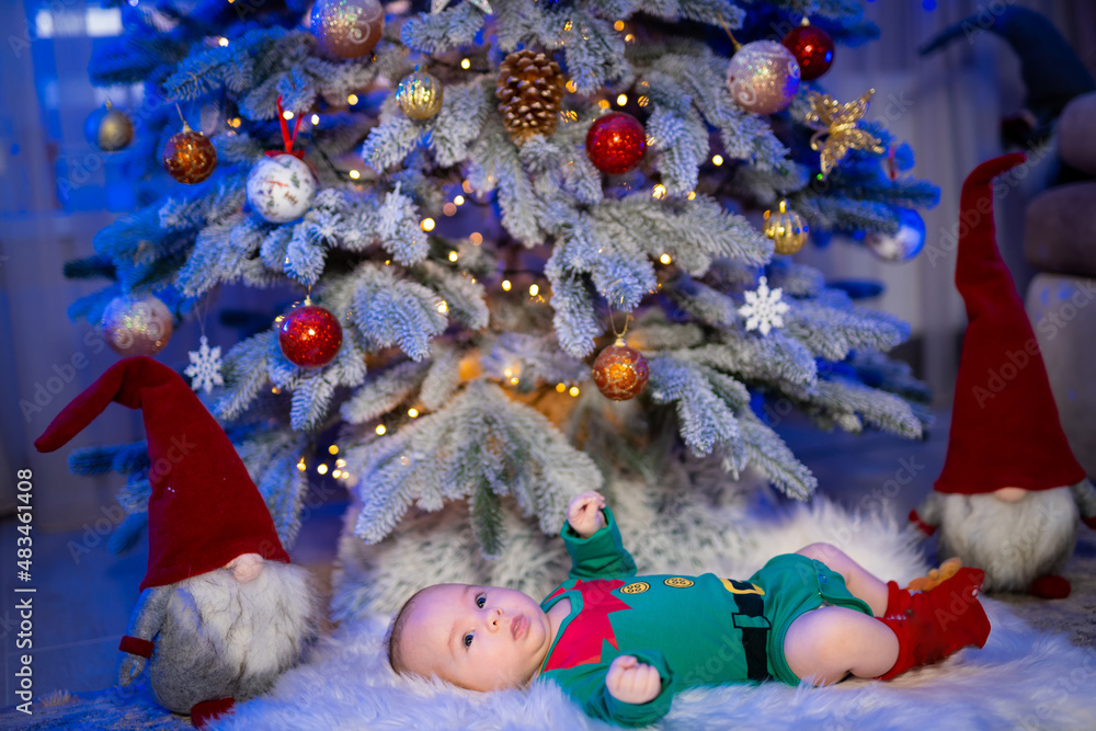 Sweet baby lying on the floor in a costume of elf. Baby near the fur tree at the new year. Side view