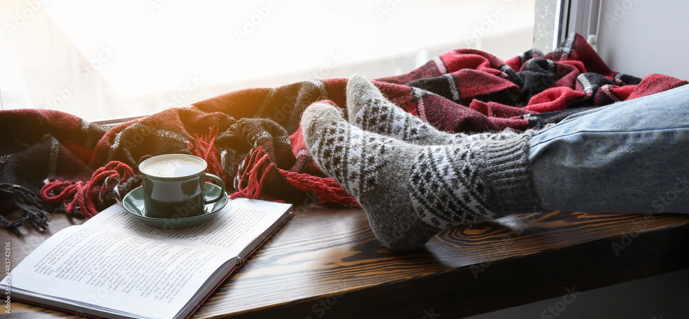 Female legs in warm socks with cup of coffee and book near window at home. Concept of heating season