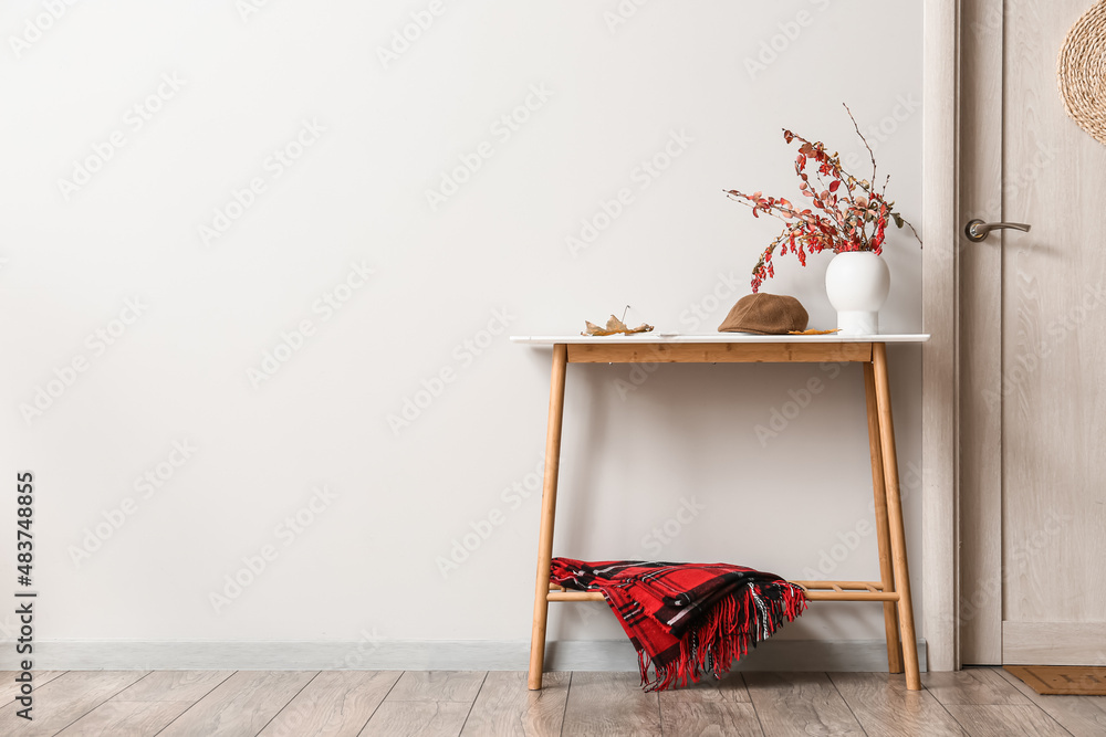 Vase with autumn branches and hat on table near light wall