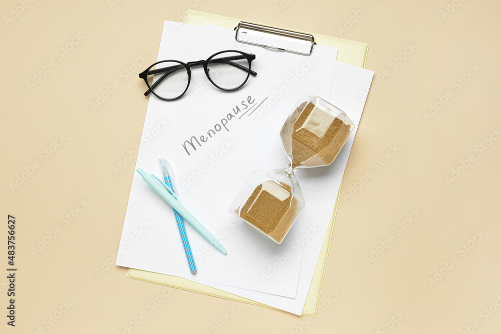 Clipboard with word MENOPAUSE, eyeglasses, pens and hourglass on beige background