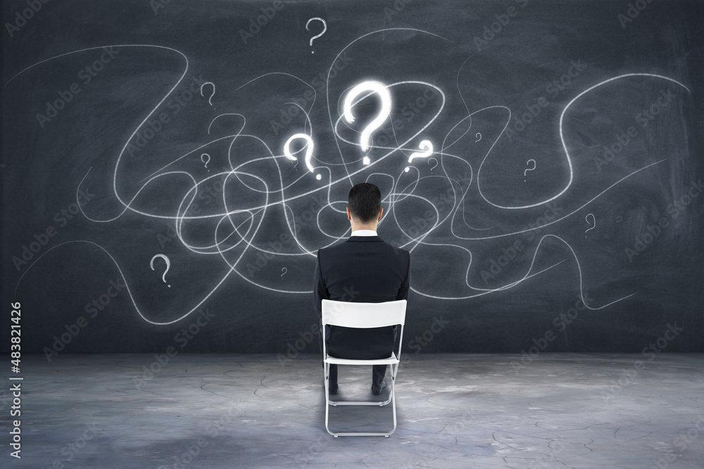 Back view of young european businessman sitting on chair on concrete wall background with drawn ques