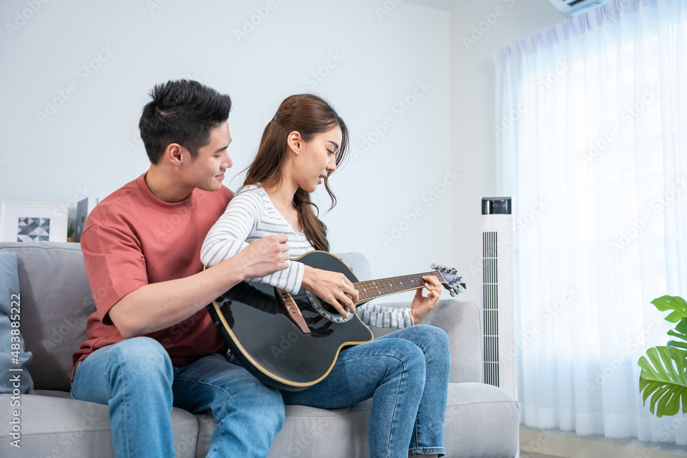 Asian young loving couple play guitar together in living room at home. 