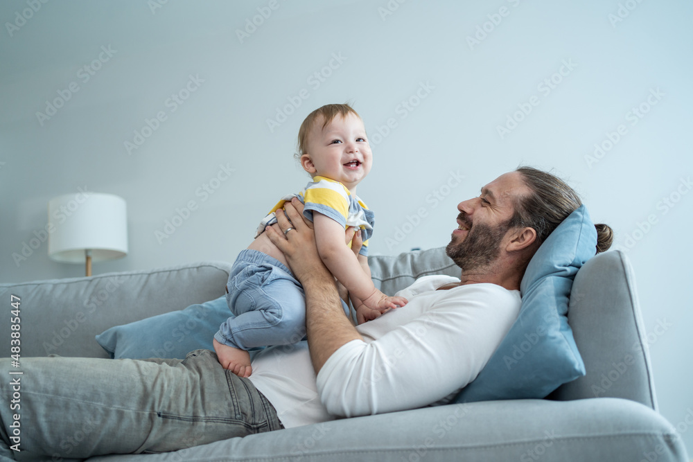 Caucasian loving dad play with baby boy child on sofa in living room.