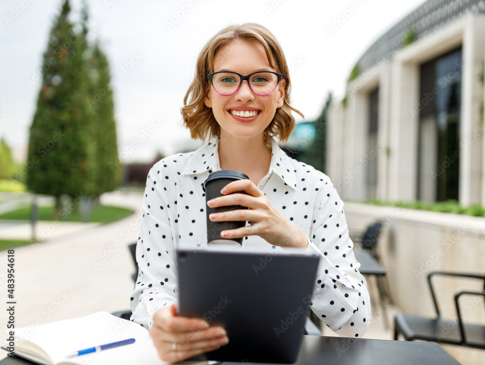 Beautiful young woman entrepreneur having cup of coffee while holding meeting online outside in cafe