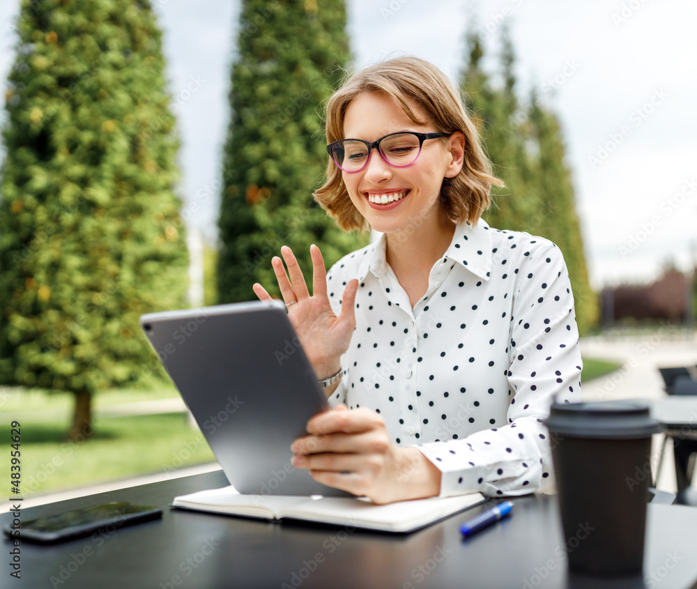 Happy young female teacher in earphones sitting in front of tablet outside having lesson online