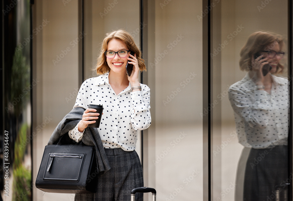 Young successful business lady talking on mobile phone while standing on city street with suitcase