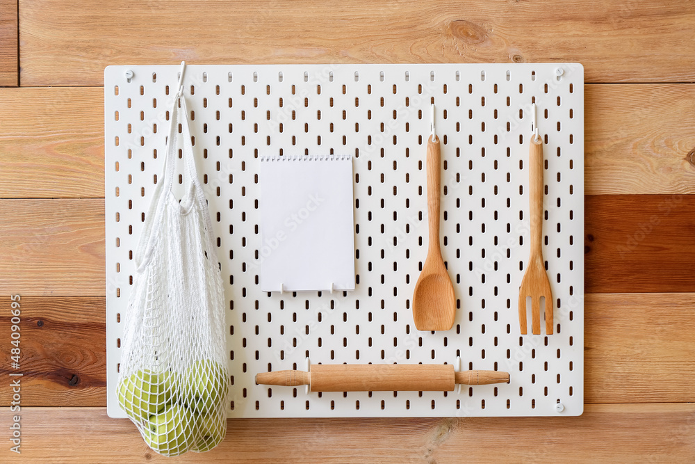 Pegboard with kitchen utensils, string bag with apples and notebook on wooden wall