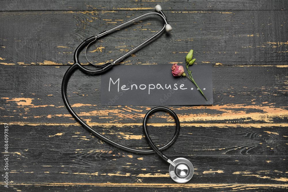 深色木质背景上写着MENOPAUSE字样的纸、干玫瑰和听诊器