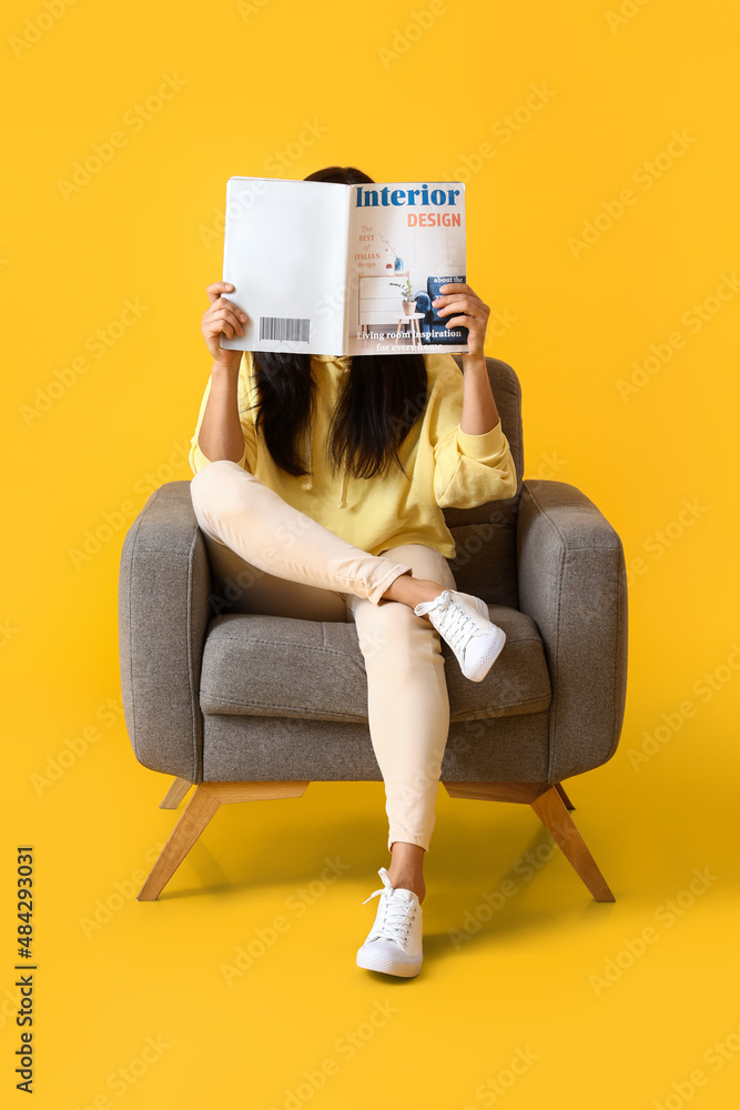 Beautiful young woman reading magazine in soft armchair on yellow background