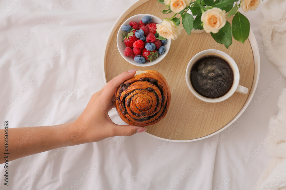 Woman taking bun from tray with breakfast on bed, closeup