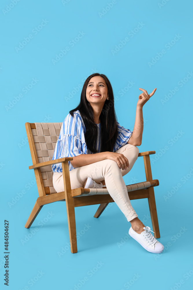 Beautiful young woman in wooden armchair pointing at something on blue background