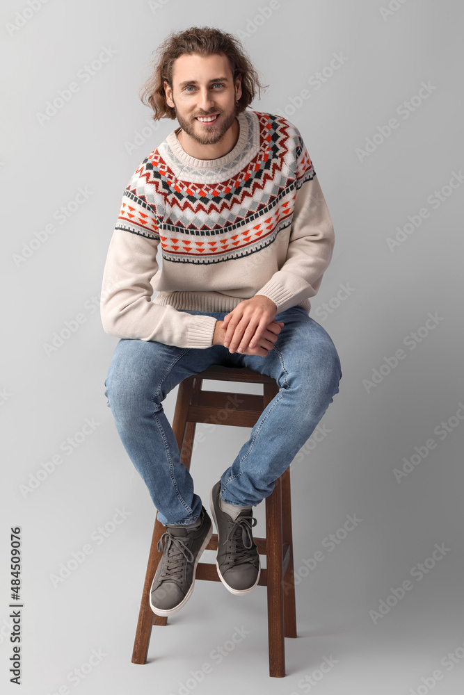 Handsome young man in knitted sweater sitting on chair against grey background