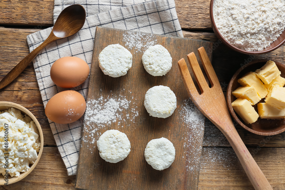 Board with uncooked cottage cheese pancakes and ingredients on wooden background