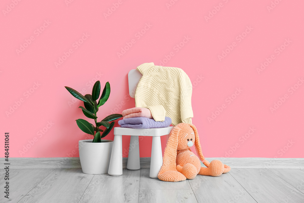 Toy, houseplant and chair with knitted sweaters for children near pink wall