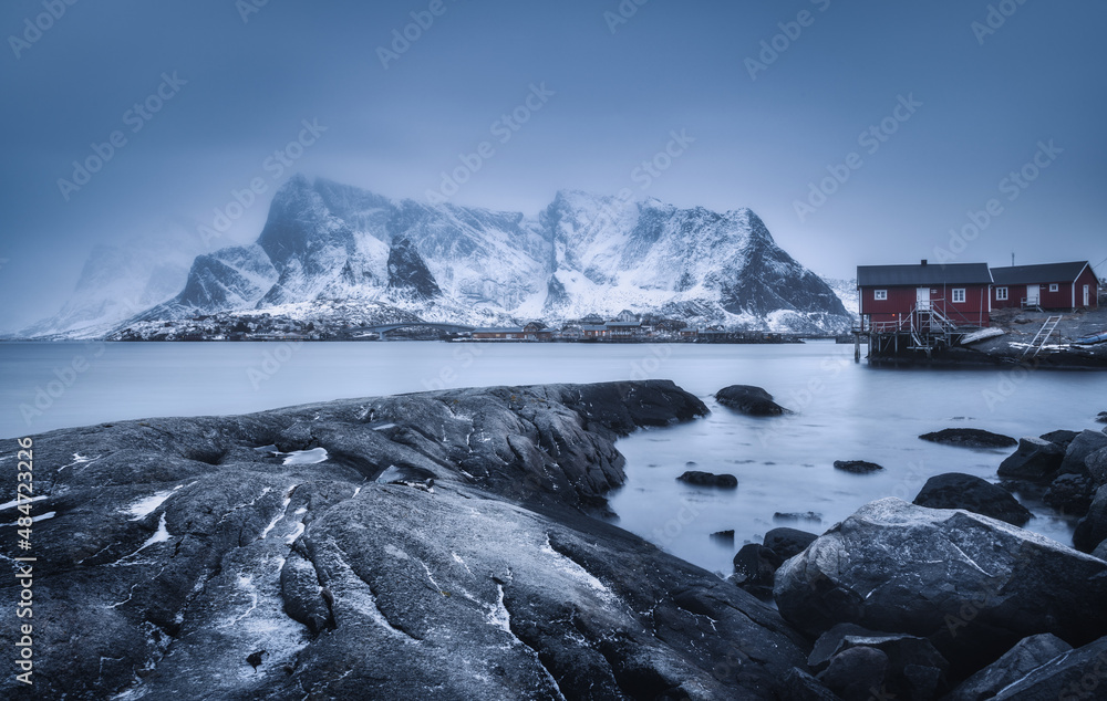 Beach with stones in blurred water, red rorbu and snowy mountains in fog at dusk in winter. Sea coas