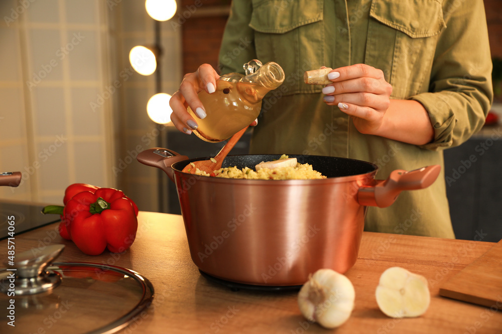 Woman adding oil to cooking pot with pilaf on wooden table in kitchen