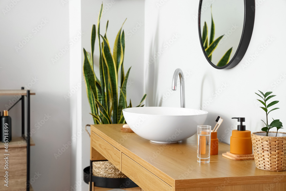 Wooden table with sink and bath supplies near light wall in room