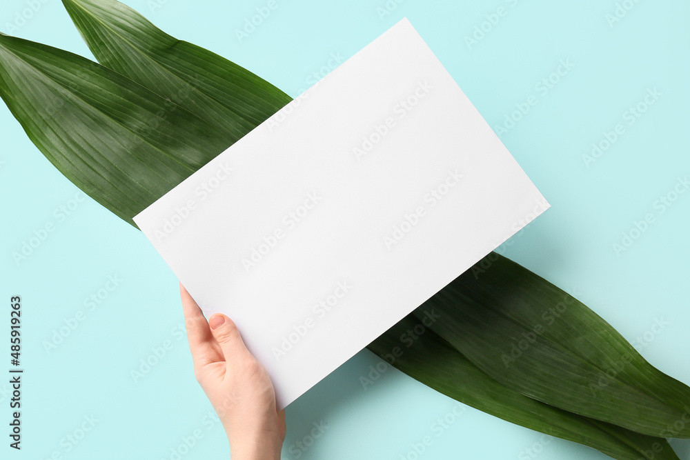 Female hand with blank card and plant leaves on color background, closeup