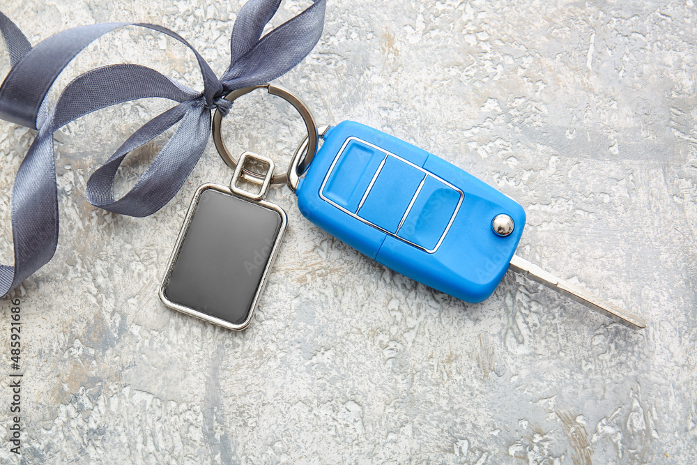 Ribbon with car key and stylish square keychain on light background