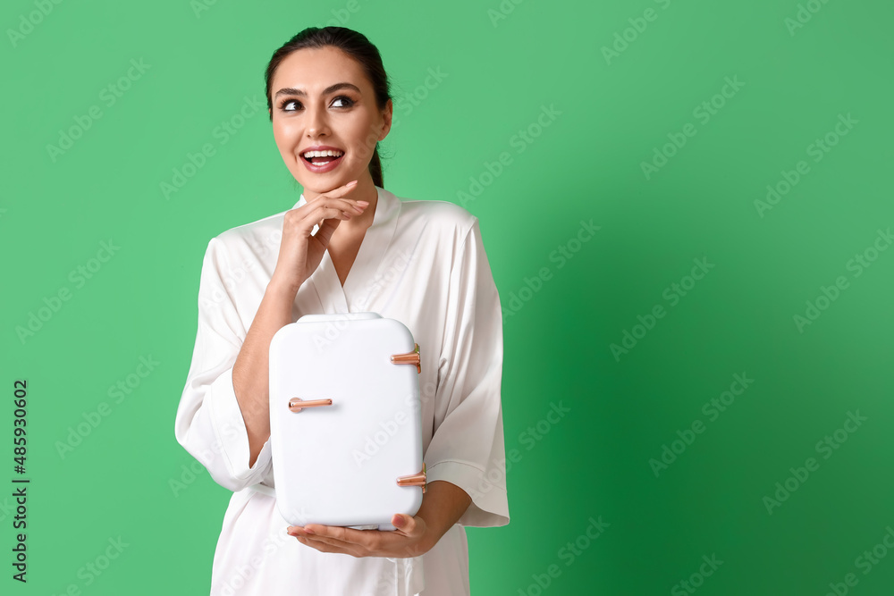 Happy young woman with small cosmetic refrigerator on green background