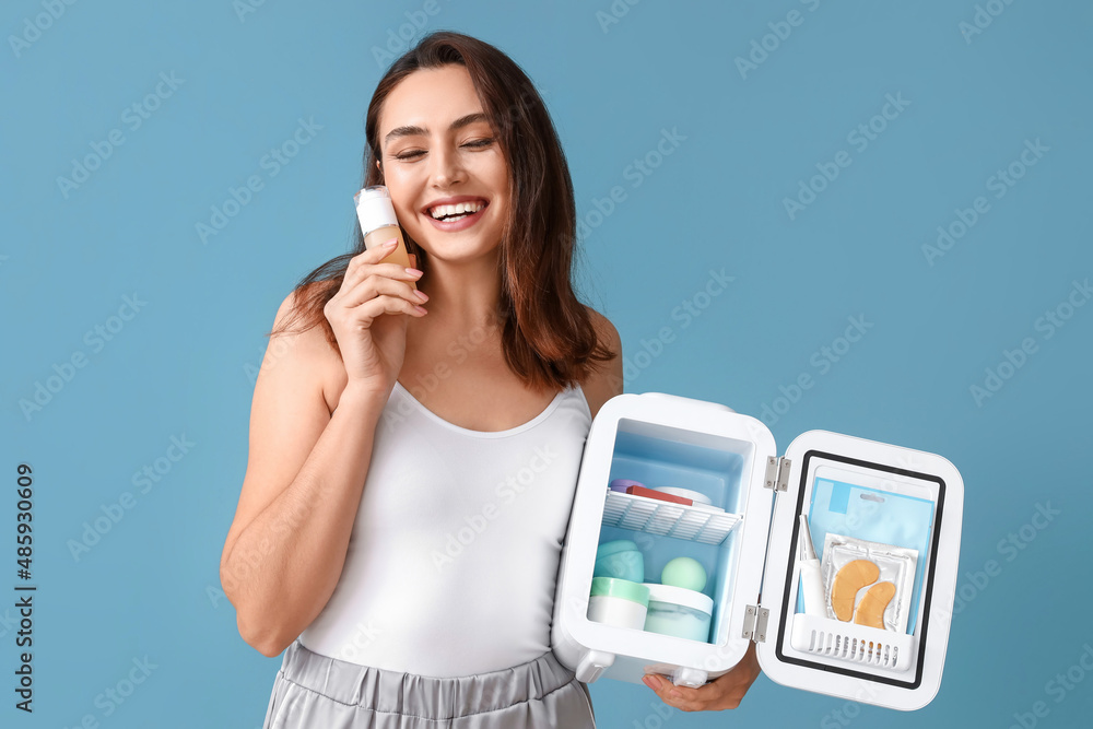 Happy young woman with open cosmetic refrigerator and products on blue background