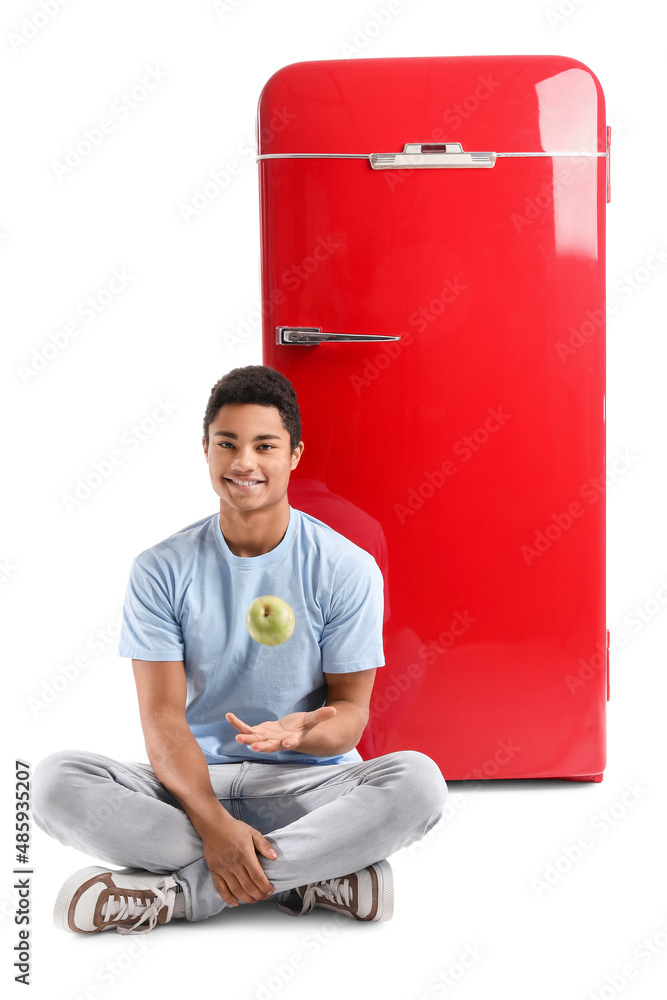 African-American man sitting with apple near stylish red fridge on white background