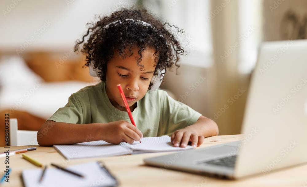 Smiling african american child school boy in headphones studying online on laptop at home