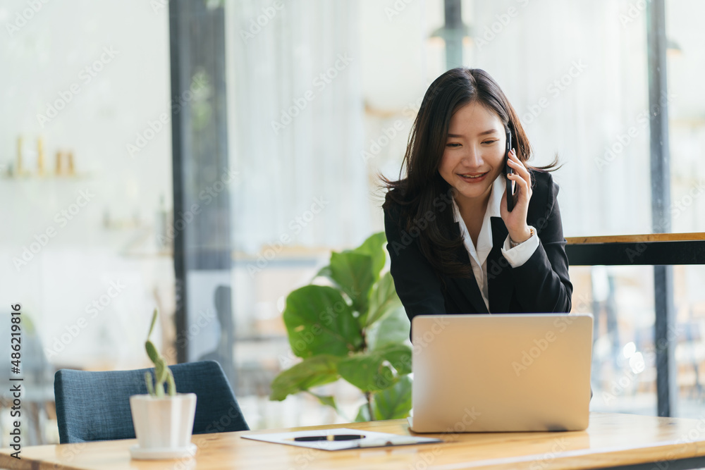 Happy smiling Asian business woman working on laptop at office, using smart phone. Businesswoman sit