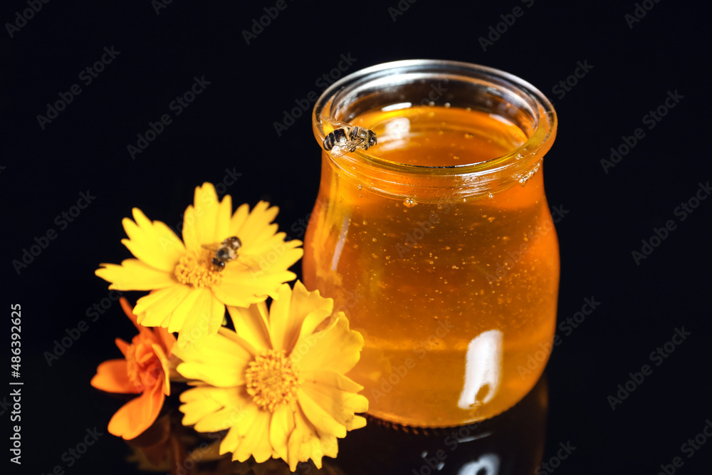 Glass jar with honey, marigold flowers and bees on black background