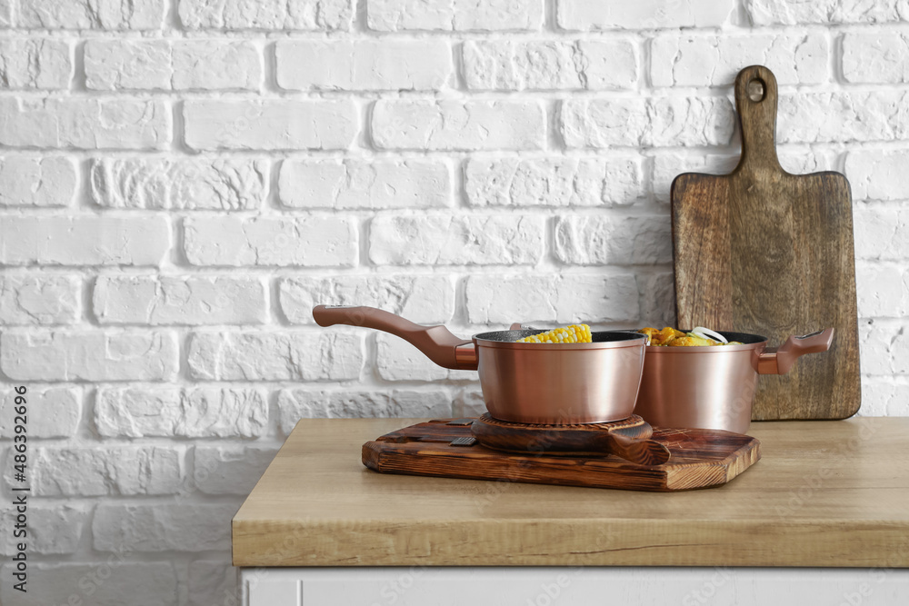 Sauce pan, cooking pot with prepared food and wooden boards on table near white brick wall