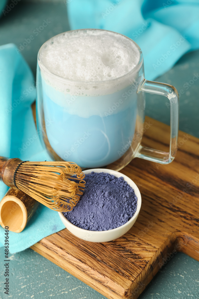 Wooden board with cup of blue matcha latte powder and chasen on color background, closeup