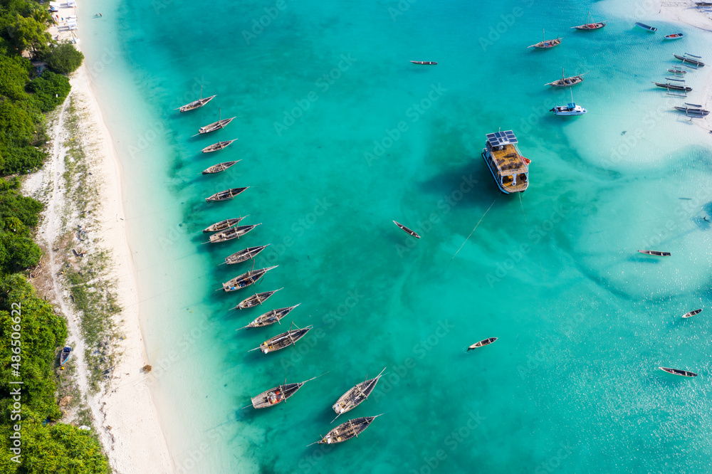 Many boats on the ocean coast and thin strip of sandy beach, palm trees. View from above on the turq