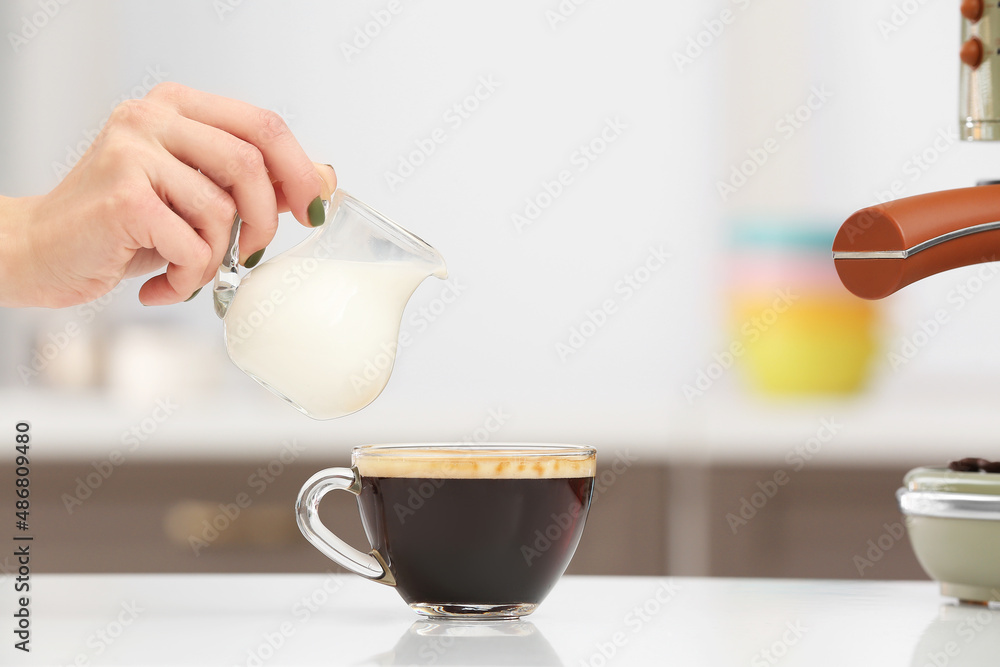 Woman pouring milk into cup of fresh coffee on table in kitchen