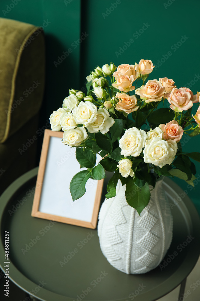 Vase with bouquet of beautiful fresh roses and blank frame on table in room