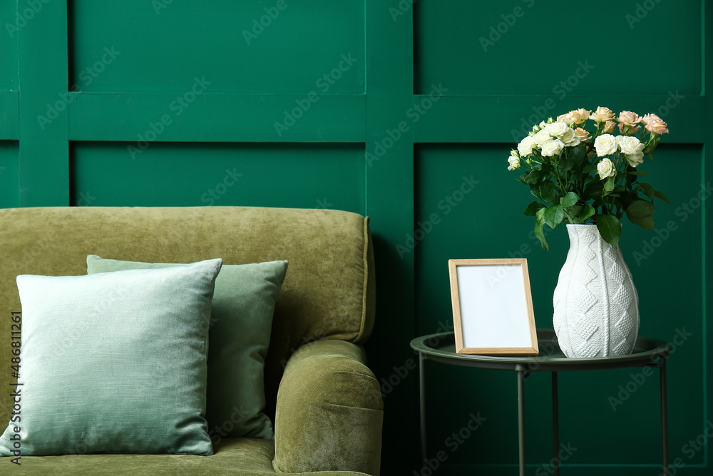 Vase with bouquet of beautiful fresh roses on table and comfortable sofa near green wall