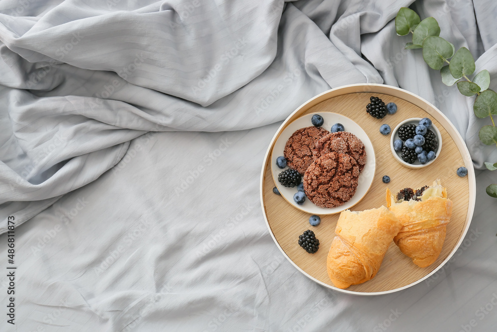 Tray with delicious croissant, cookies, berries and eucalyptus branch on bed