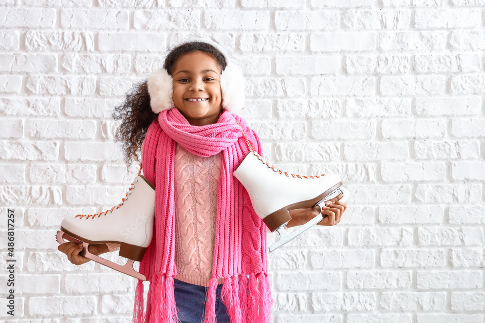 Little African-American girl with ice skates on white brick background