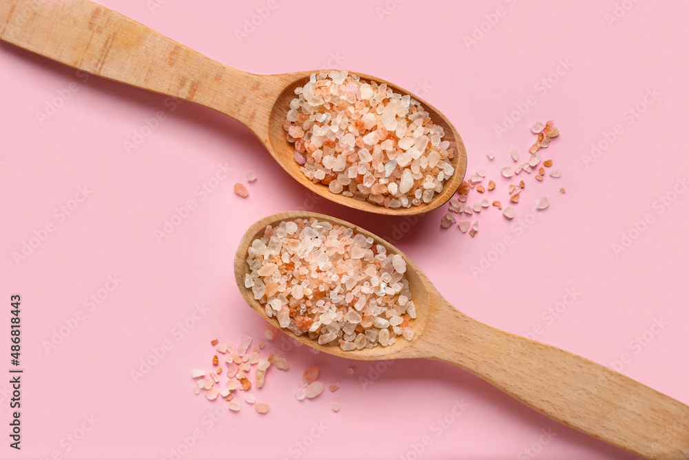 Wooden spoons with sea salt on pink background, closeup