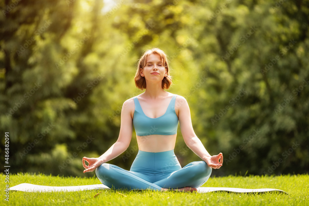 Happy adorable sporty girl performing yoga while sitting on mat in lotus position in nature