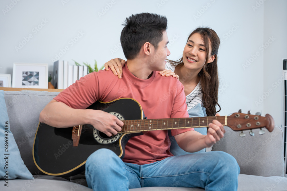 Asian young loving couple play guitar together in living room at home. 