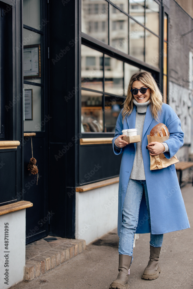 Joyful adult woman, holding cups of takeaway coffee, and pastry, while walking.