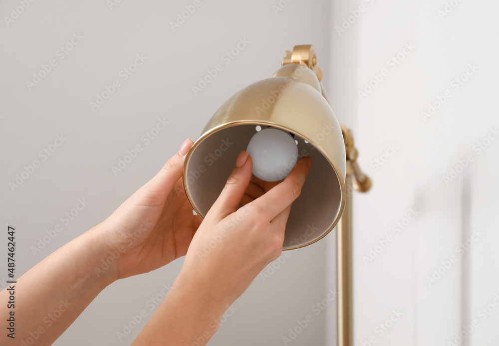 Woman changing light bulb in golden lamp near light wall, closeup